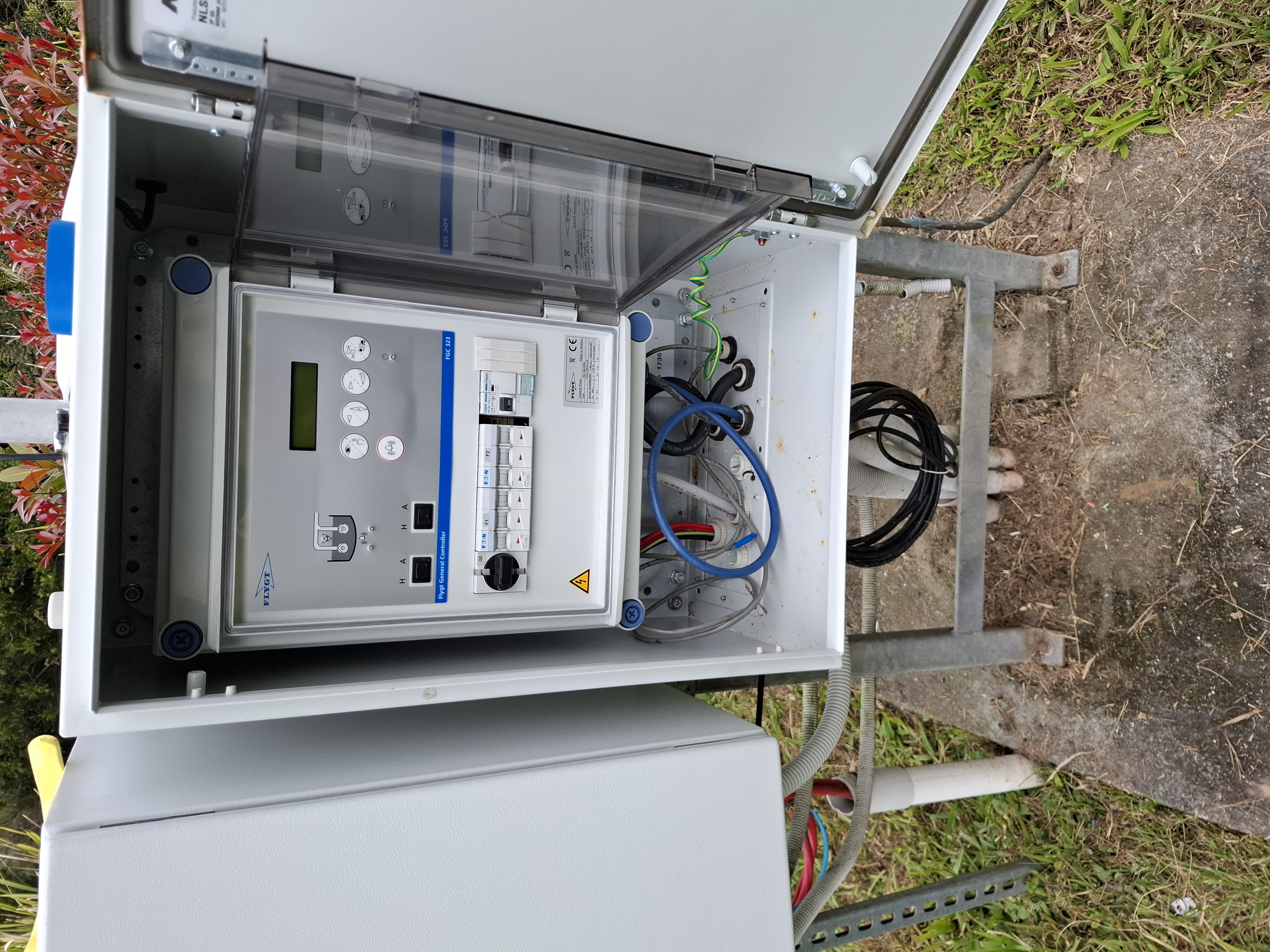 New Goto Engineering telemetry enclosure installed alongside the existing Flygt pump controller cabinet on a stainless stand, antenna mast above